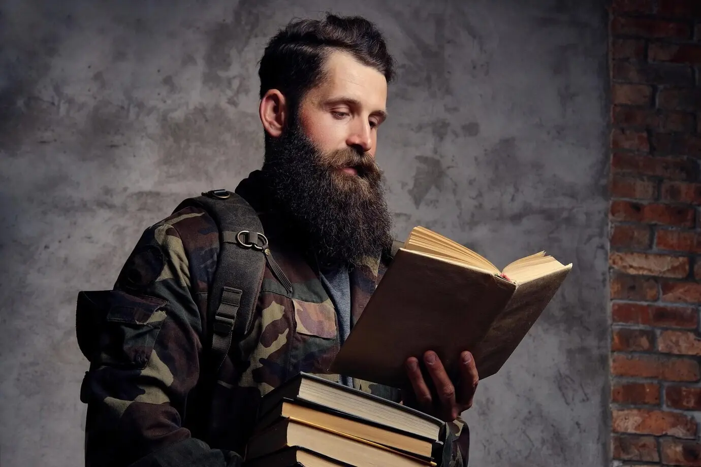 Ein gutaussehender, bärtiger Hipster in einer trendigen Camouflage-Jacke steht im Studio, hält einen Bücherstapel in den Händen und liest, isoliert vor dunklem Hintergrund.